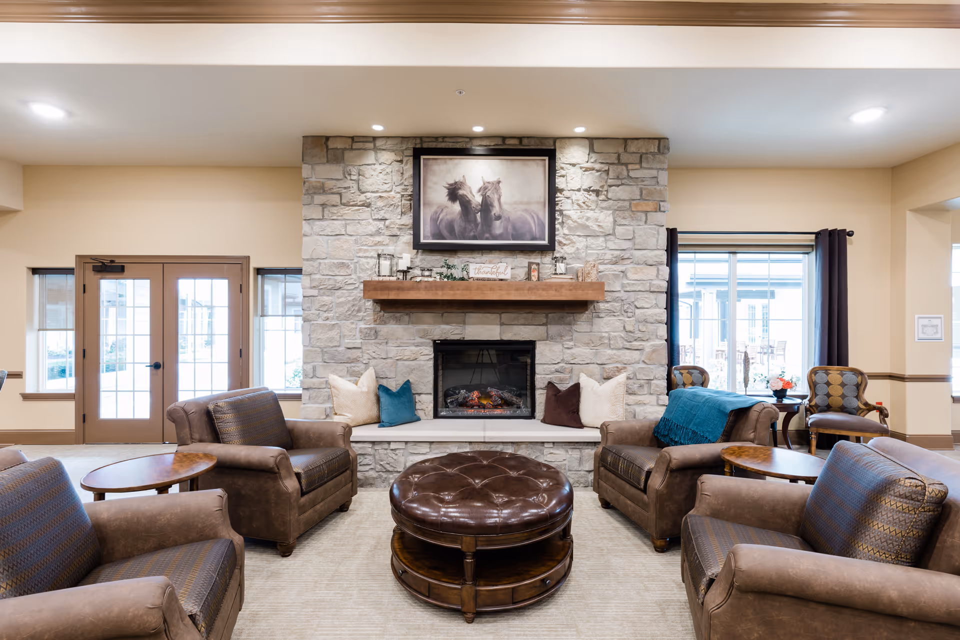 A cozy living room area with a stone fireplace in the center, topped with a framed picture of two horses. The room features four brown leather armchairs arranged around a round, tufted leather ottoman. There are windows and glass doors on either side of the fireplace, allowing natural light to enter. The walls are painted a warm beige color, and the room has a calm, inviting atmosphere.