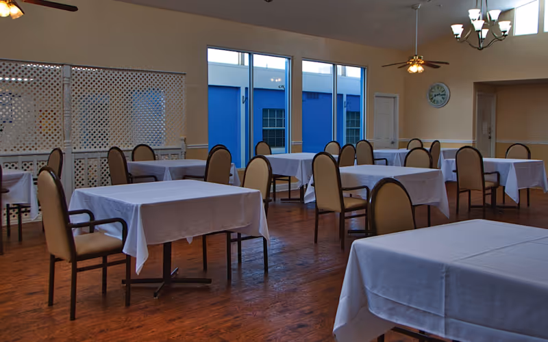 A dining room with multiple tables covered in white tablecloths and beige cushioned chairs arranged around them. The room has wooden flooring, large windows showing a blue exterior wall, ceiling fans, and a chandelier. A clock is mounted on the wall.