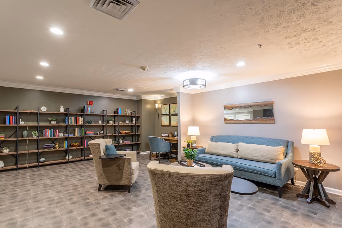 A cozy living room area in a senior living facility featuring a blue sofa with two beige cushions, two beige armchairs, a round coffee table with a small plant, and two side tables with lamps. The room has a large bookshelf filled with books and decorative items against a green wall, a small desk with a blue chair in the corner, and a rectangular mirror with a wooden frame hanging above the sofa. The ceiling has recessed lighting and a modern circular light fixture.