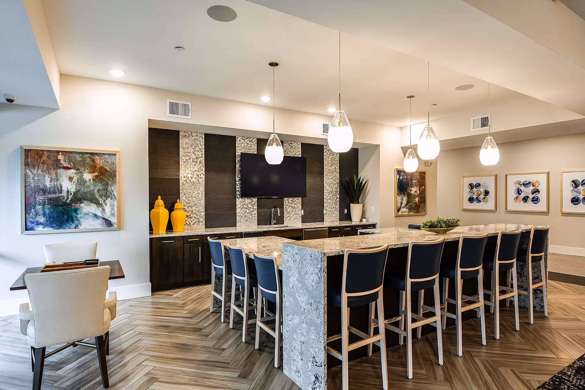 Modern communal dining area with a large marble island surrounded by high-back chairs. The back wall features dark wood cabinetry, a mounted flat-screen TV, a small sink, and decorative yellow vases. Pendant lights hang from the ceiling, and abstract artwork adorns the walls. The floor has a herringbone wood pattern.