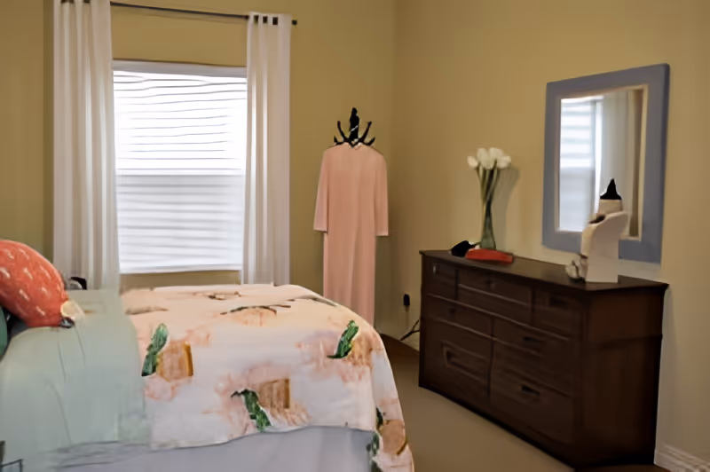 A bedroom with a bed covered in a floral patterned blanket and pillows, a window with white curtains, a wooden dresser with a mirror above it, a vase with white flowers, and a pink dress on a mannequin stand in the corner.
