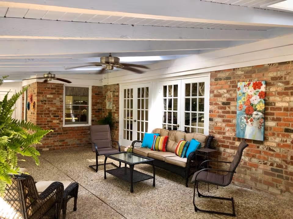 Covered outdoor patio area with brick walls, white wooden ceiling beams, and two ceiling fans. The space is furnished with a cushioned wicker sofa with colorful pillows, two chairs, and a glass-top coffee table. There is a colorful floral painting on the brick wall and green plants adding to the decor.