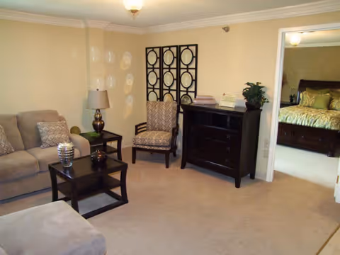 A cozy living room with beige walls and carpet, featuring a beige sofa with patterned cushions, a dark wood coffee table with decorative items, a matching side table with a lamp, an upholstered armchair, and a dark wood dresser with a plant and books. A doorway leads to a bedroom with a bed and green bedding.