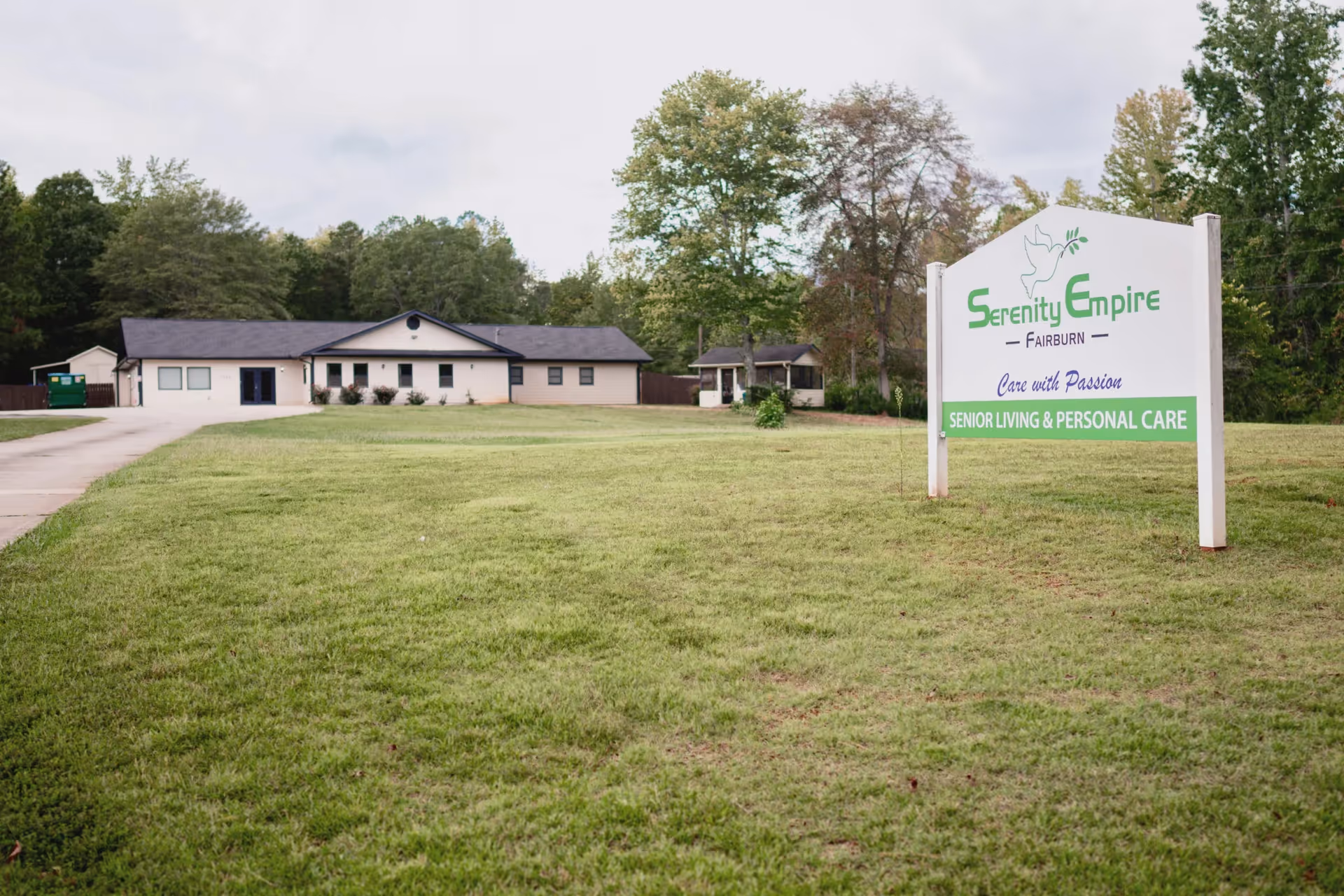 A single-story building with a dark roof and light-colored walls set behind a large grassy lawn. To the right, there is a white sign with green and purple text that reads 'Serenity Empire Fairburn Care with Passion Senior Living & Personal Care.' Trees and a cloudy sky are visible in the background.