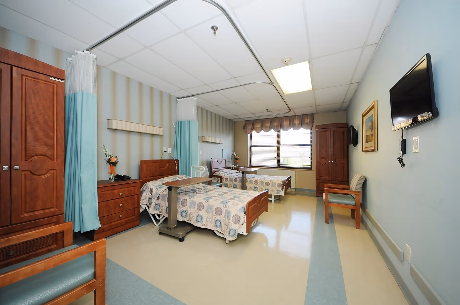A bright and clean senior living bedroom with two hospital-style beds, each with a patterned bedspread. The room has light blue walls, a large window with blinds and a valance, wooden wardrobes, a chest of drawers with a vase of flowers, a wall-mounted TV, and a wooden chair. Privacy curtains hang from ceiling tracks around each bed.