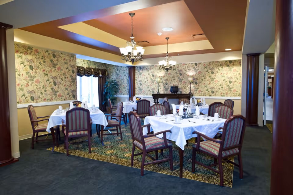 A dining room with several tables covered in white tablecloths, each set with cups, napkins, and utensils. The room features floral wallpaper, striped upholstered chairs, chandeliers hanging from a recessed ceiling, and a carpeted floor with a patterned rug under the tables.