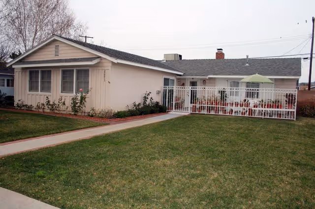 Single-story residential building with beige exterior walls, a gray shingled roof, and multiple windows. There is a fenced patio area with white metal fencing, outdoor chairs, and a green umbrella. The building is surrounded by a well-maintained lawn and a concrete walkway leading to the entrance.