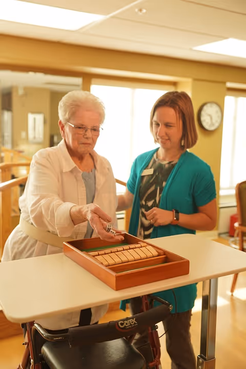 An elderly woman with a walker is playing a wooden dice game on a table, assisted by a younger woman wearing a teal cardigan and a name badge, in a well-lit room with large windows and a clock on the wall.