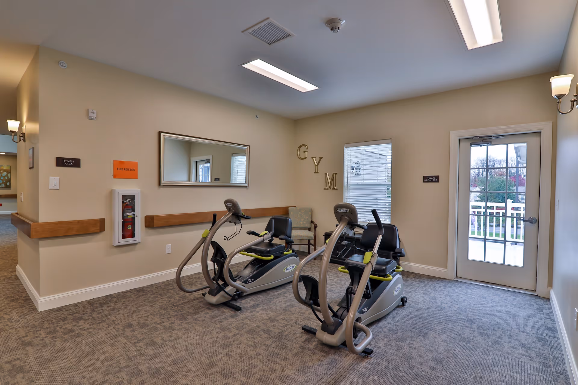 A small fitness area inside Heather Glen Senior Living with two stationary exercise bikes, a chair, a large mirror on the wall, and a glass door leading outside. The walls are beige, and there is a carpeted floor. The word 'GYM' is displayed on the wall near the window.