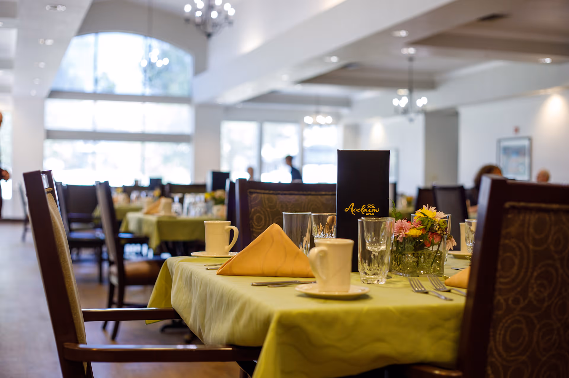 A dining room in Acclaim Living facility with tables covered in green tablecloths, set with cups, glasses, utensils, napkins, and small flower arrangements. The room has large windows allowing natural light and chandeliers hanging from the ceiling.