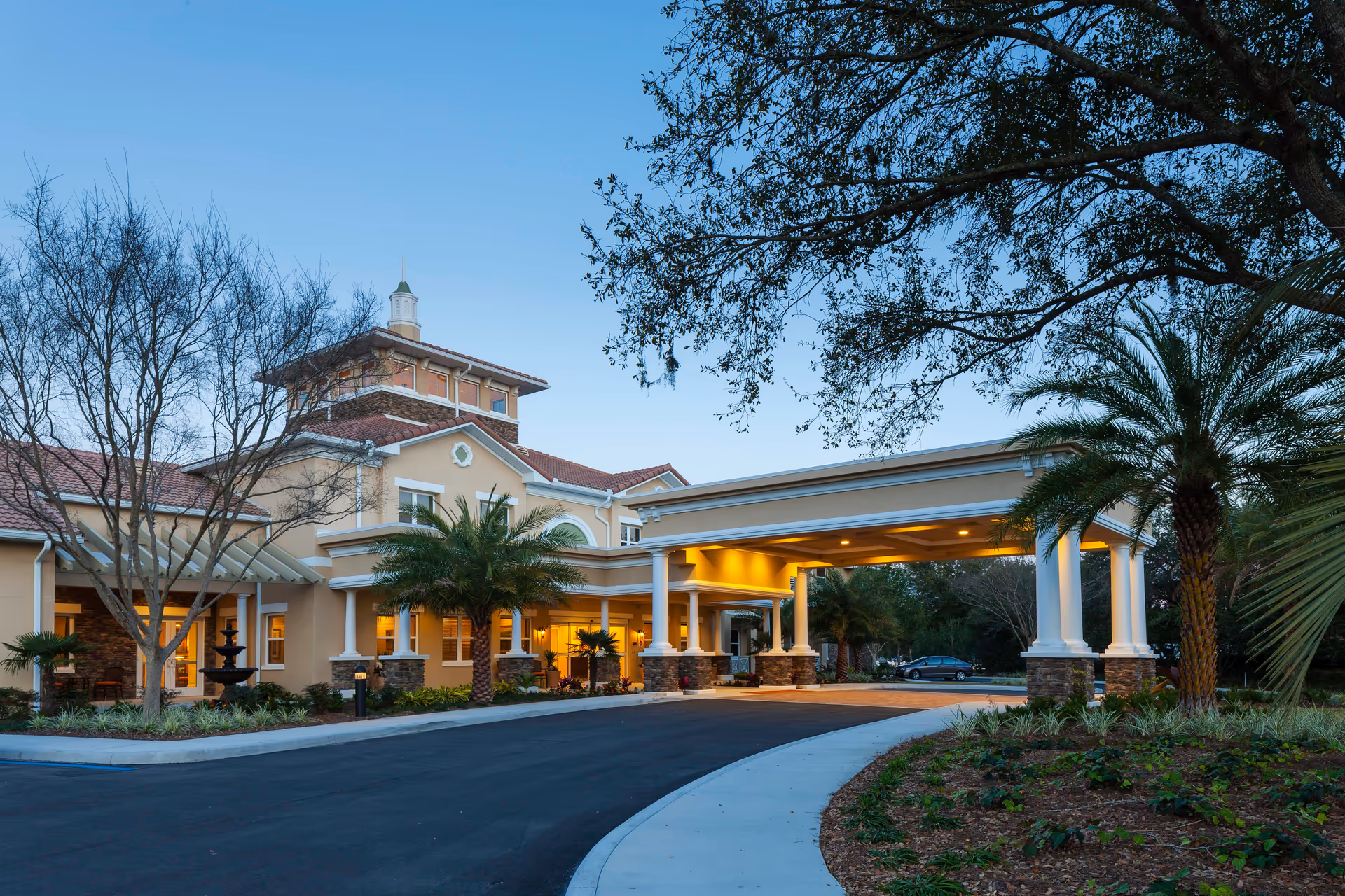 Exterior view of HarborChase of Villages Crossing senior living facility at dusk, showing the main entrance with a covered driveway, palm trees, landscaped garden, and warm lighting illuminating the building.