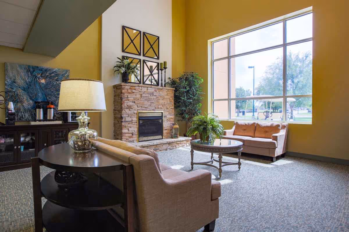 A bright living room area with large windows letting in natural light. The room features two beige sofas, a round glass coffee table with a green plant, a stone fireplace with decorative items on the mantel, and a side table with a lamp. There is also a cabinet with a coffee machine and a large painting on the wall.