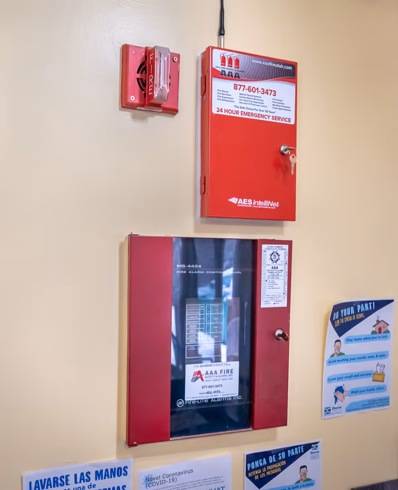 Wall-mounted fire alarm control panel and fire alarm pull station in a facility, with informational posters about COVID-19 and hygiene in Spanish and English displayed below and to the side.