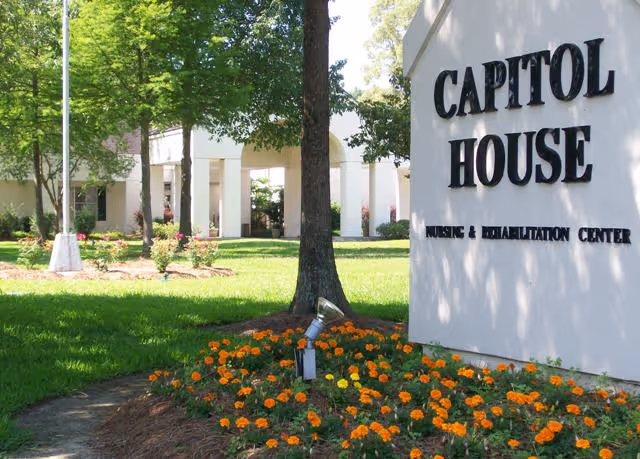 Outdoor view of Capitol House Nursing & Rehabilitation Center with a white sign displaying the facility name, surrounded by green grass, orange flowers, and trees. The building entrance with white columns is visible in the background.