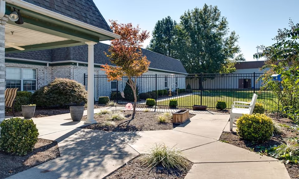 Outdoor courtyard area at The Arbors at WestRidge Place Senior Living featuring a paved walkway, small trees, bushes, and a white bench. The courtyard is enclosed by a black metal fence with a building and clear blue sky in the background.