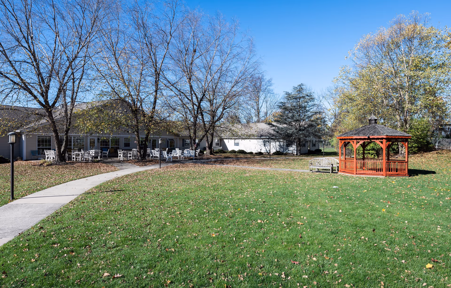 Outdoor area of a senior living facility with a green lawn, a red wooden gazebo, a wooden bench, and a paved walkway leading to a building with multiple white chairs and tables outside. Trees with sparse leaves surround the area under a clear blue sky.