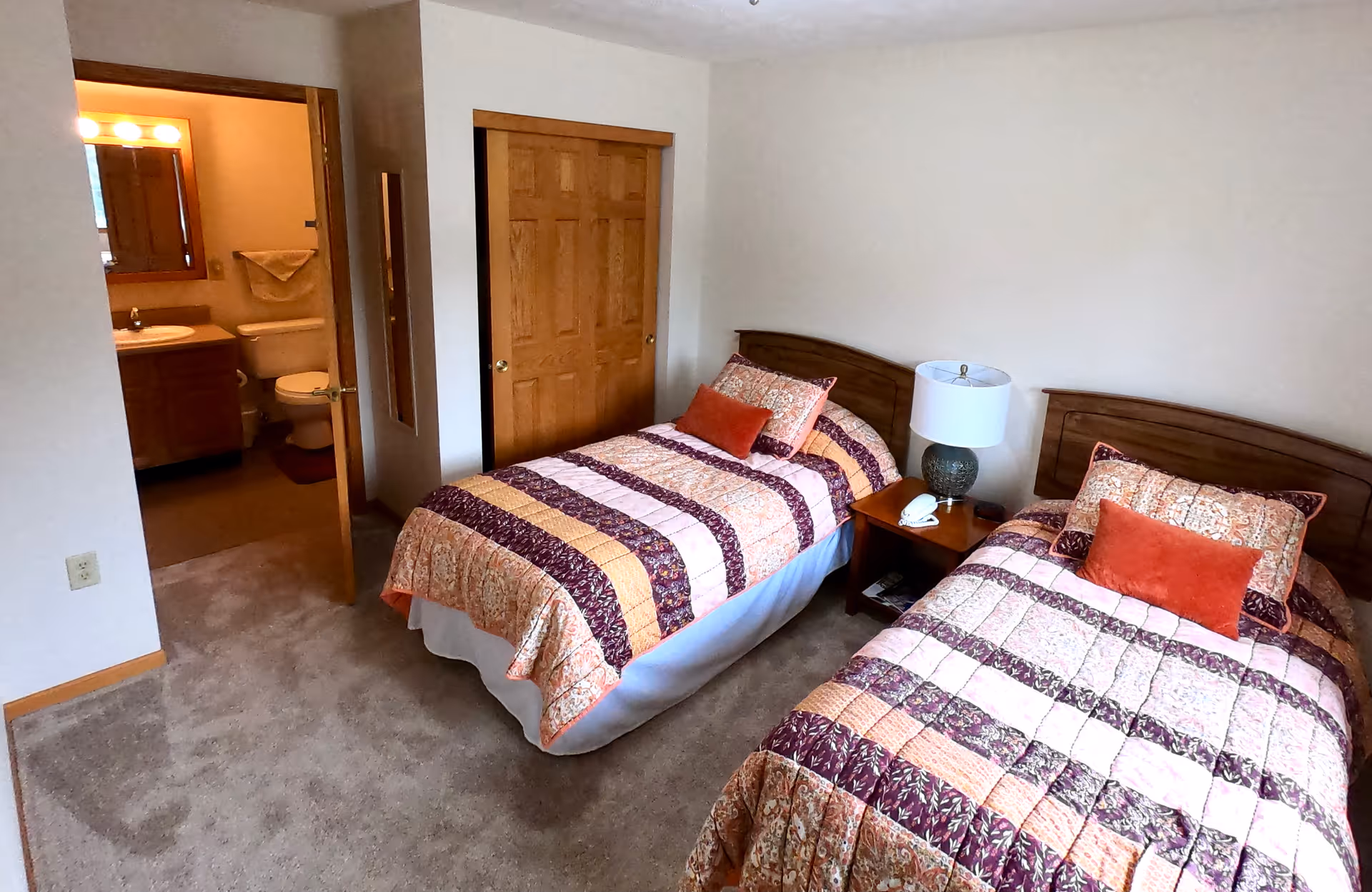A bedroom with two twin beds covered in patterned quilts with orange and purple tones. Between the beds is a wooden nightstand with a lamp and a telephone. The room has beige carpet and white walls. There is a wooden closet door and an open door leading to a bathroom with a toilet, sink, and mirror visible.