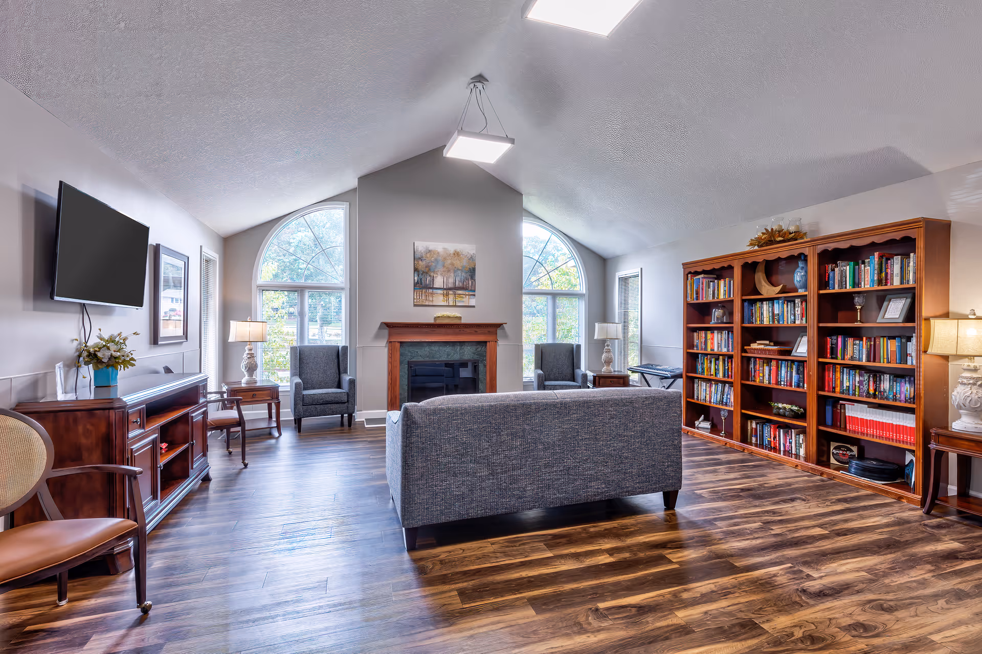 A bright and spacious living room with vaulted ceilings, two large arched windows, a fireplace with a painting above it, a gray sofa facing the fireplace, two gray armchairs, a wooden bookshelf filled with books, a wall-mounted flat-screen TV, and wooden flooring.