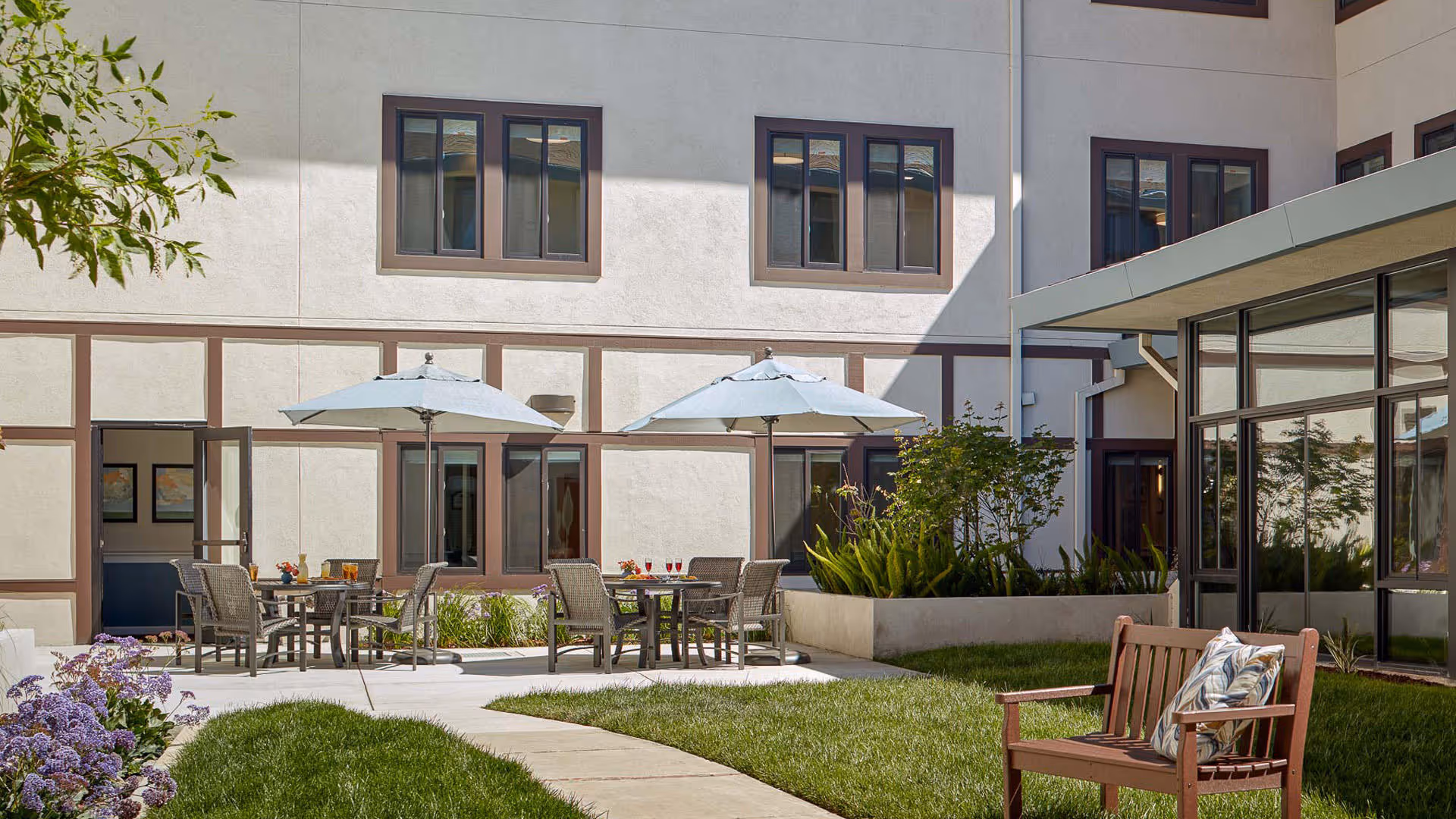 Outdoor courtyard area at Atria Park of San Mateo featuring patio tables with umbrellas, chairs, green grass, plants, and a wooden bench with a decorative pillow. The building exterior with windows and doors surrounds the courtyard.