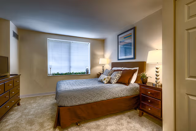 Well-lit bedroom with a made bed, two nightstands with lamps, a dresser and a window with blinds.