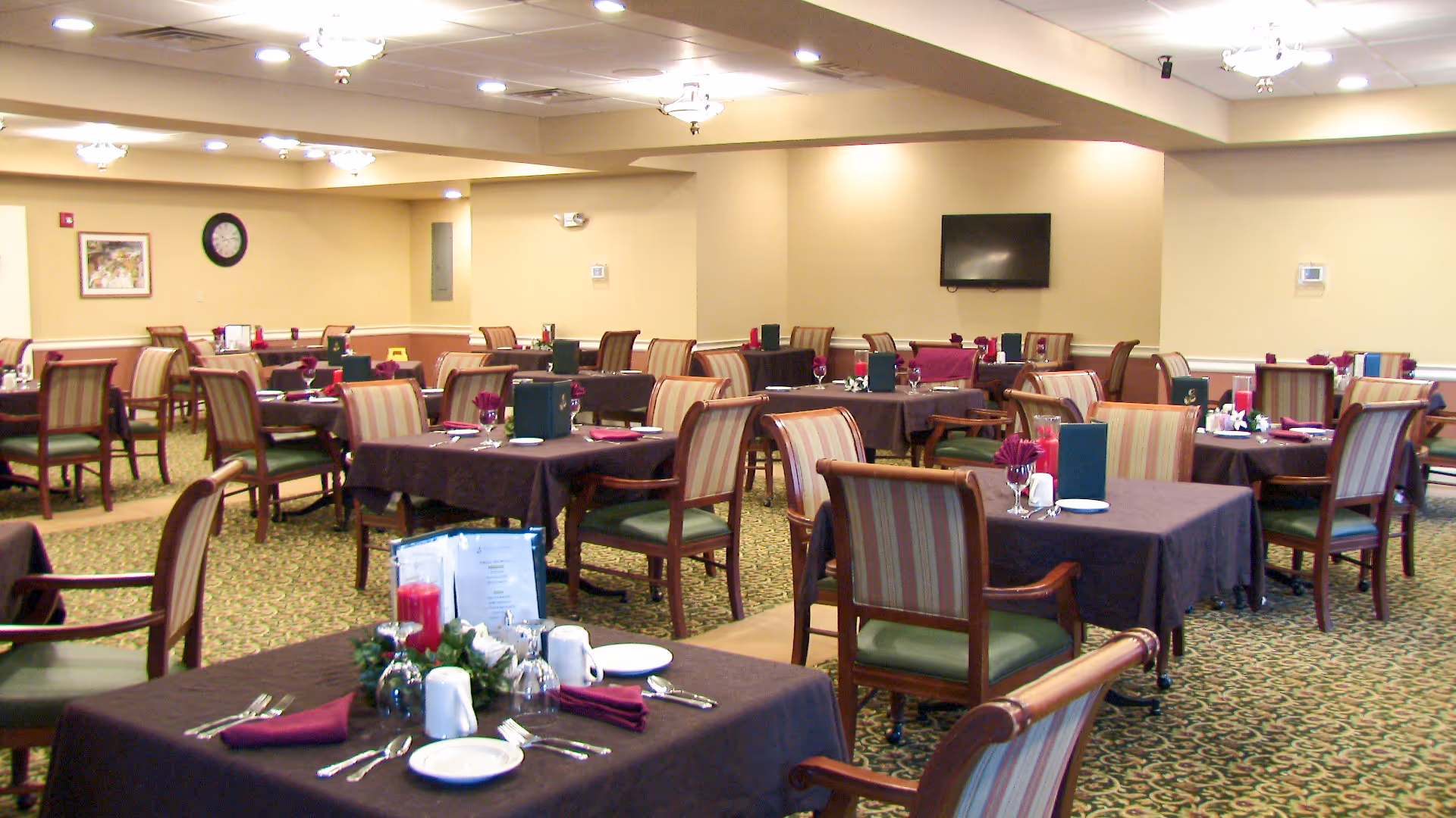 Dining room with multiple set tables, upholstered chairs, and place settings in a senior living facility.