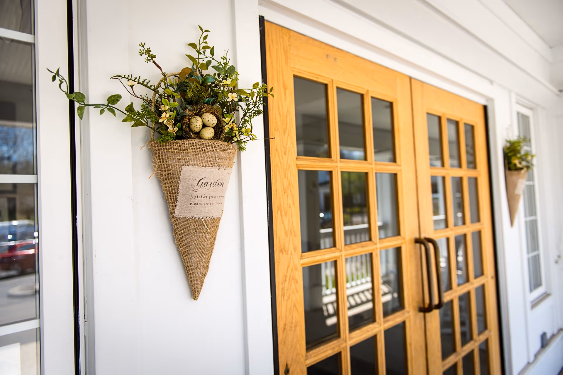 A close-up view of a decorative burlap wall hanging with artificial greenery and speckled eggs, mounted on a white wall next to a wooden double door with glass panes.