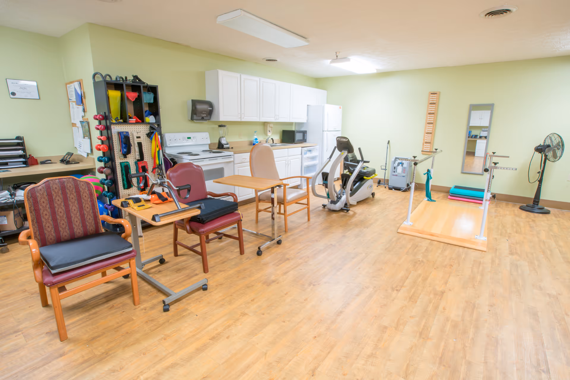 A spacious room with light green walls and wooden flooring featuring exercise equipment including a stationary bike, parallel bars, and various therapy tools. There are two chairs with small tables on wheels, a kitchen area with white cabinets, a stove, refrigerator, and a microwave. A fan and a mirror are mounted on the wall.