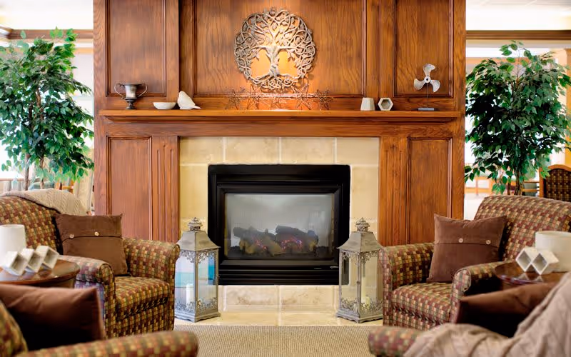 Cozy seating area with patterned armchairs facing a central fireplace set in a wood-paneled surround, flanked by plants and decorative lanterns.