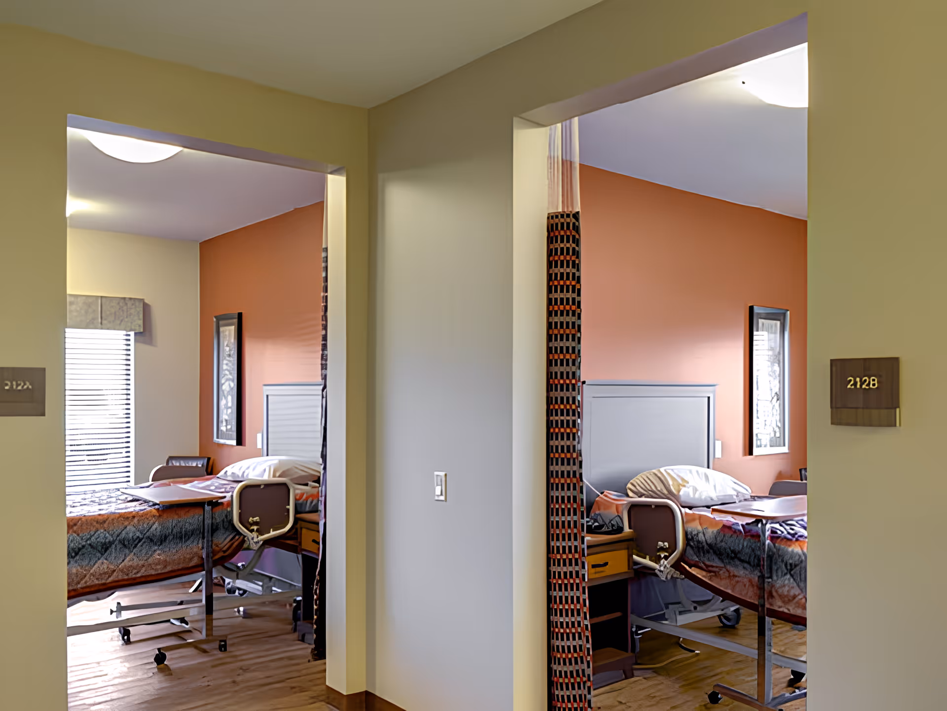 View of two adjacent patient rooms in a care facility, each with a hospital bed, bedside table, chair, and a window with blinds. The walls are painted in a warm orange color, and the floors are wooden. Room numbers 212A and 212B are visible on plaques outside each room.