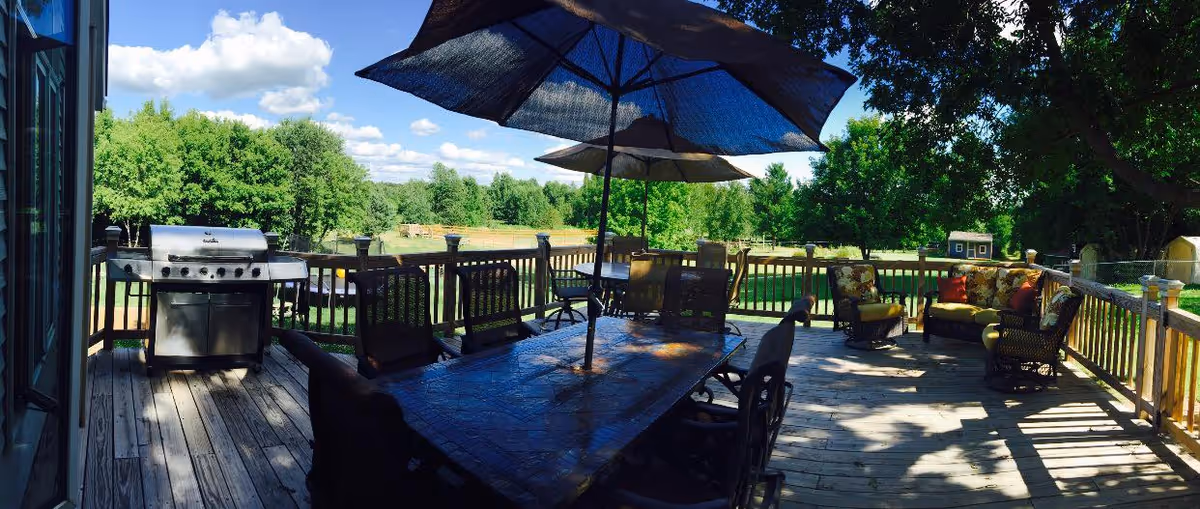 Outdoor wooden deck with a large dining table and chairs under umbrellas, a grill on the left side, and cushioned seating area with armchairs and a loveseat on the right side. The deck overlooks a green grassy area with trees and a small shed in the background under a partly cloudy blue sky.