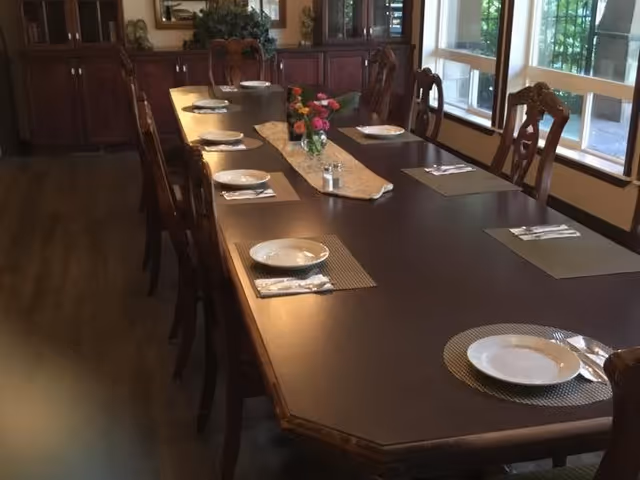 A long wooden dining table set with white plates, silverware, and placemats. A beige table runner with a small vase of colorful flowers and a salt and pepper shaker are placed in the center. The room has large windows on one side and wooden cabinets along the back wall.