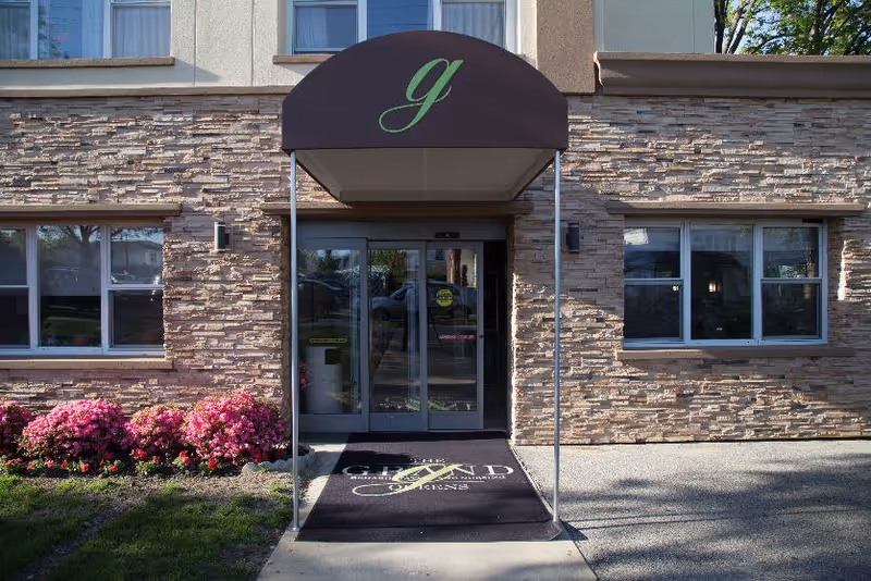 Front entrance of a brick-faced senior living building with a canopy, glass automatic doors, and flowering shrubs.