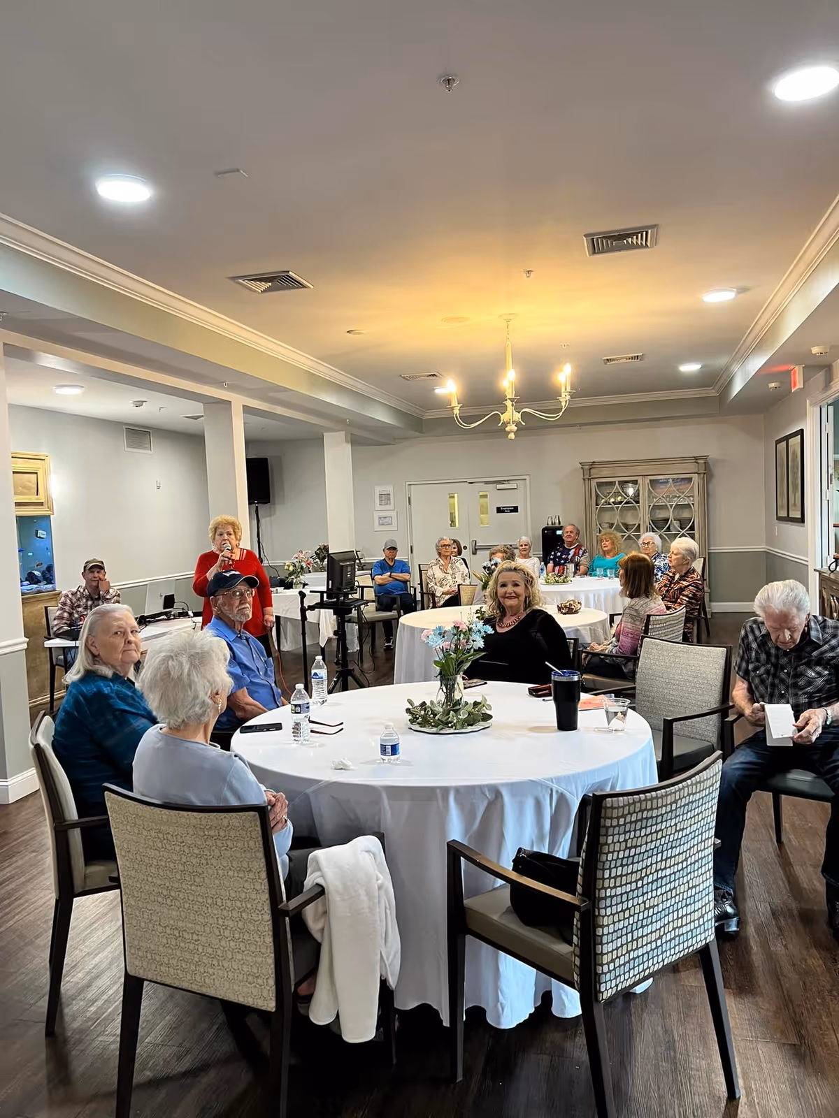 A group of senior residents seated around round tables in a bright community room with a chandelier.