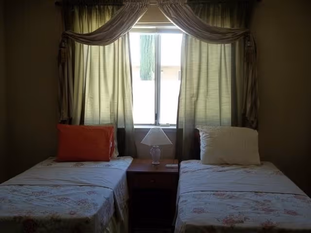 A bedroom with two single beds separated by a wooden nightstand with a lamp. Each bed has a pillow, one orange and one white. Behind the nightstand is a window with green curtains and a decorative valance.