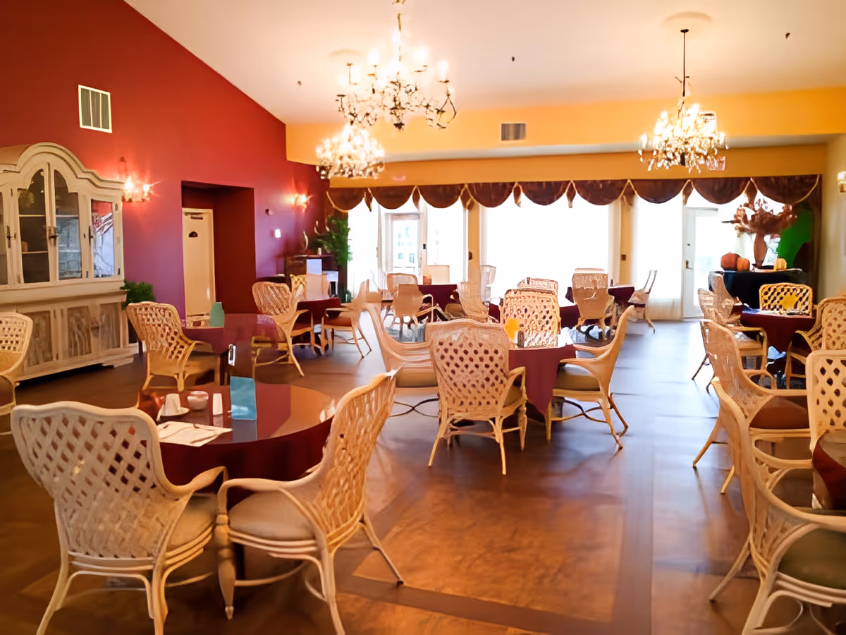 A spacious dining room with multiple round tables covered with burgundy tablecloths and surrounded by wicker chairs with cushions. The room features a red accent wall on the left, large windows with draped valances letting in natural light, chandeliers hanging from the ceiling, and a white cabinet against the red wall.