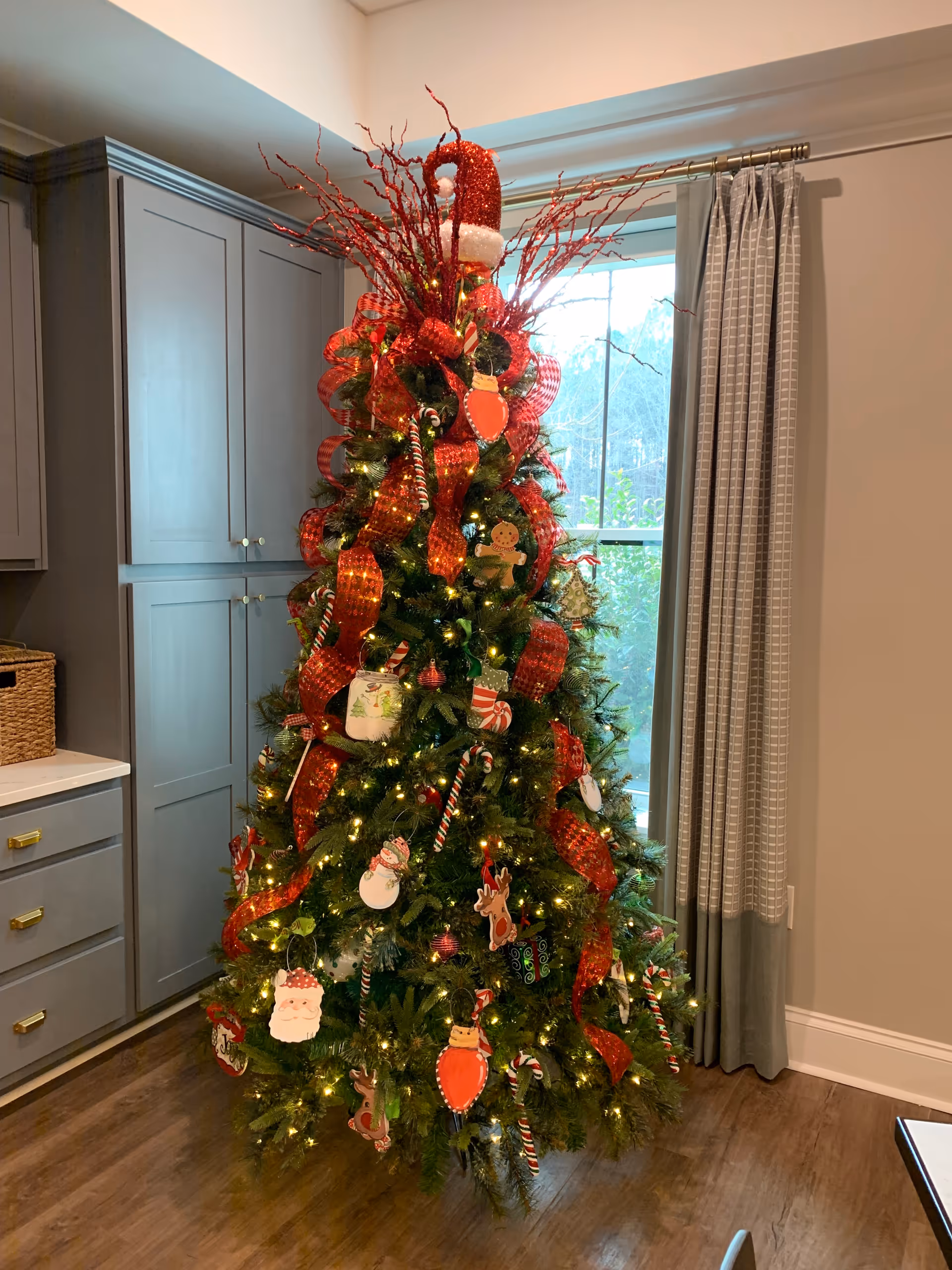 A decorated Christmas tree with red ribbons and ornaments stands in a room next to built-in cabinets and a window with curtains.