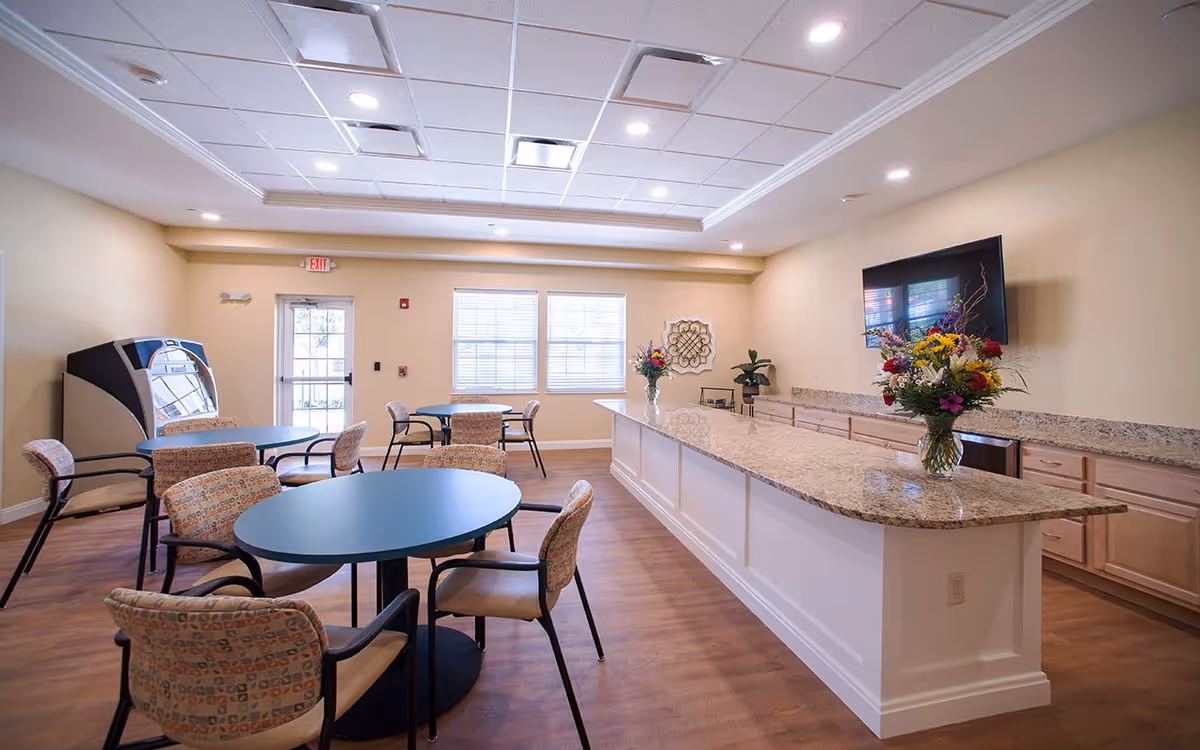 Bright communal dining room with round tables and chairs, a long granite service counter with flower arrangements, a wall-mounted TV, and windows.