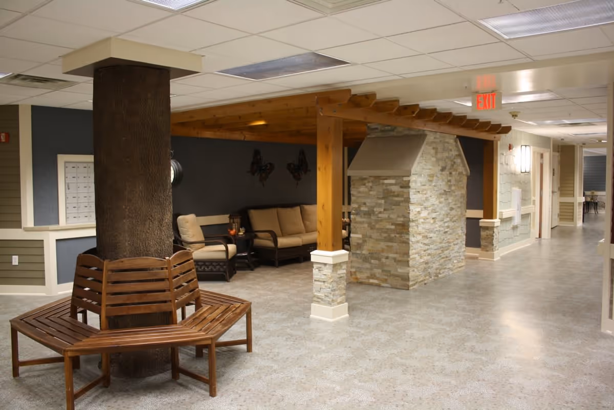 Indoor common area with a wooden bench surrounding a large tree trunk, a stone pillar with a wooden pergola above, and cushioned seating in the background. The space has a tiled floor, neutral-colored walls, and ceiling lights.