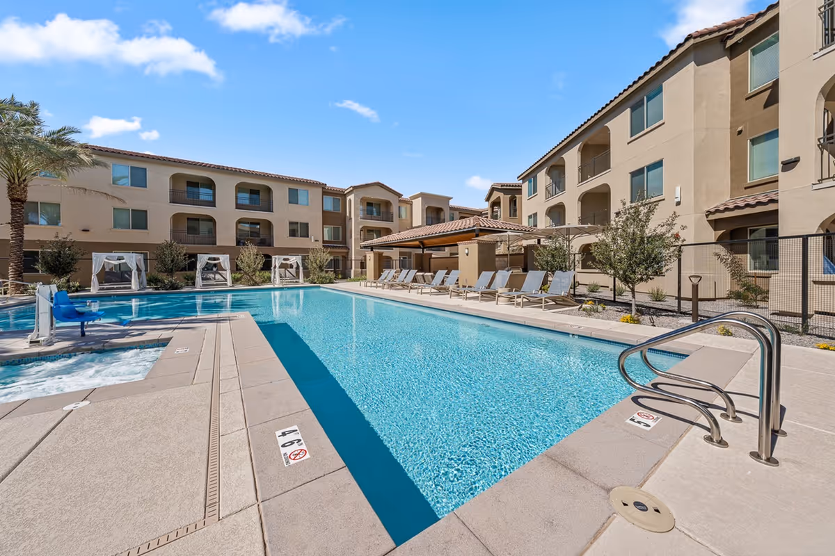 Outdoor swimming pool area at Album Marana facility with clear blue water, surrounded by lounge chairs, cabanas, palm trees, and a three-story beige building under a blue sky with some clouds.