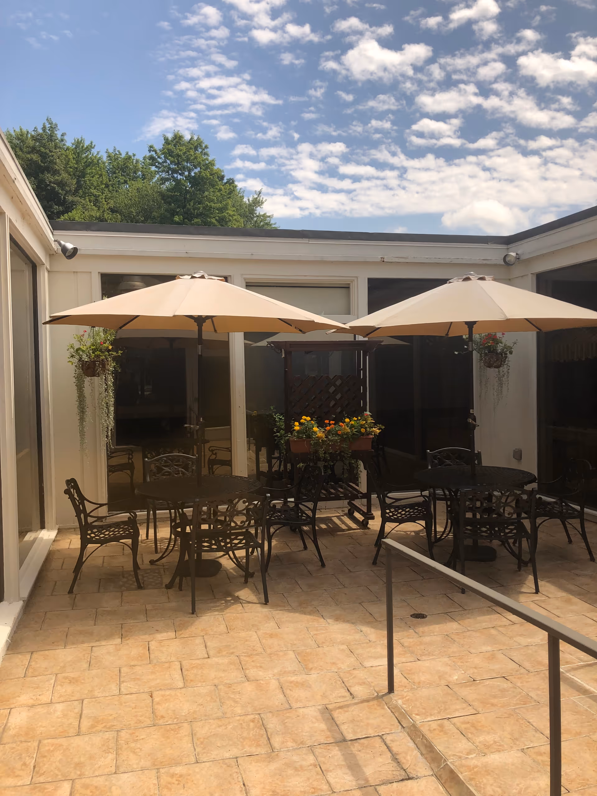 Outdoor patio area with two round metal tables, each shaded by a large beige umbrella. There are several metal chairs around the tables. Hanging flower pots with green plants are attached to the walls, and a planter with colorful flowers is placed near the center. The patio is surrounded by windows and doors, with a clear blue sky and trees visible in the background.
