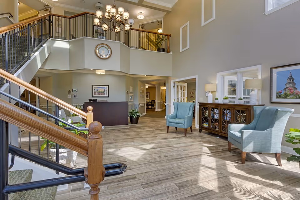 Bright and spacious senior living facility lobby with wooden flooring, a staircase with wooden handrails, two light blue armchairs, a wooden sideboard with lamps and small plants, a reception desk, a large chandelier, and a wall clock above the reception area.