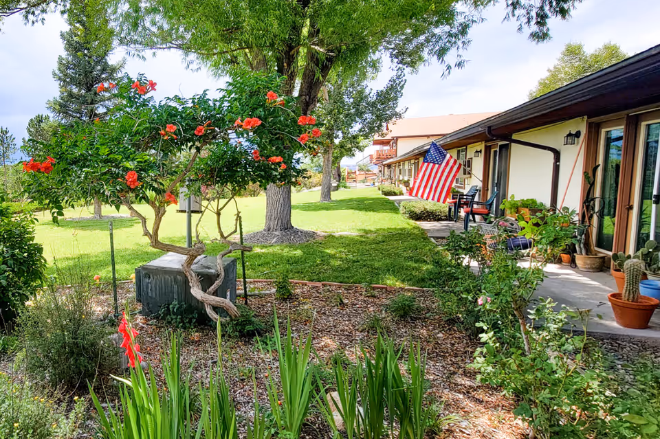 Outdoor garden area with green grass, flowering plants, and trees next to a building with a porch. The porch has potted plants, chairs, and an American flag hanging. The sky is partly cloudy.