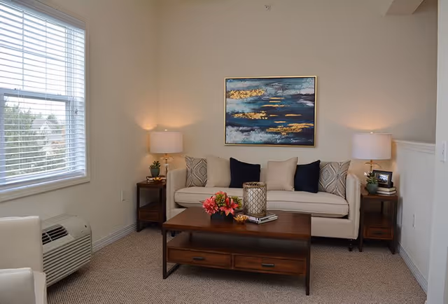 A cozy living room with a beige sofa adorned with patterned and solid cushions, flanked by two side tables each holding a lamp and decorative items. A wooden coffee table with flowers and books sits in front of the sofa. A large abstract painting with blue and gold tones hangs on the wall above the sofa. A window with white blinds allows natural light into the room, and a beige carpet covers the floor.
