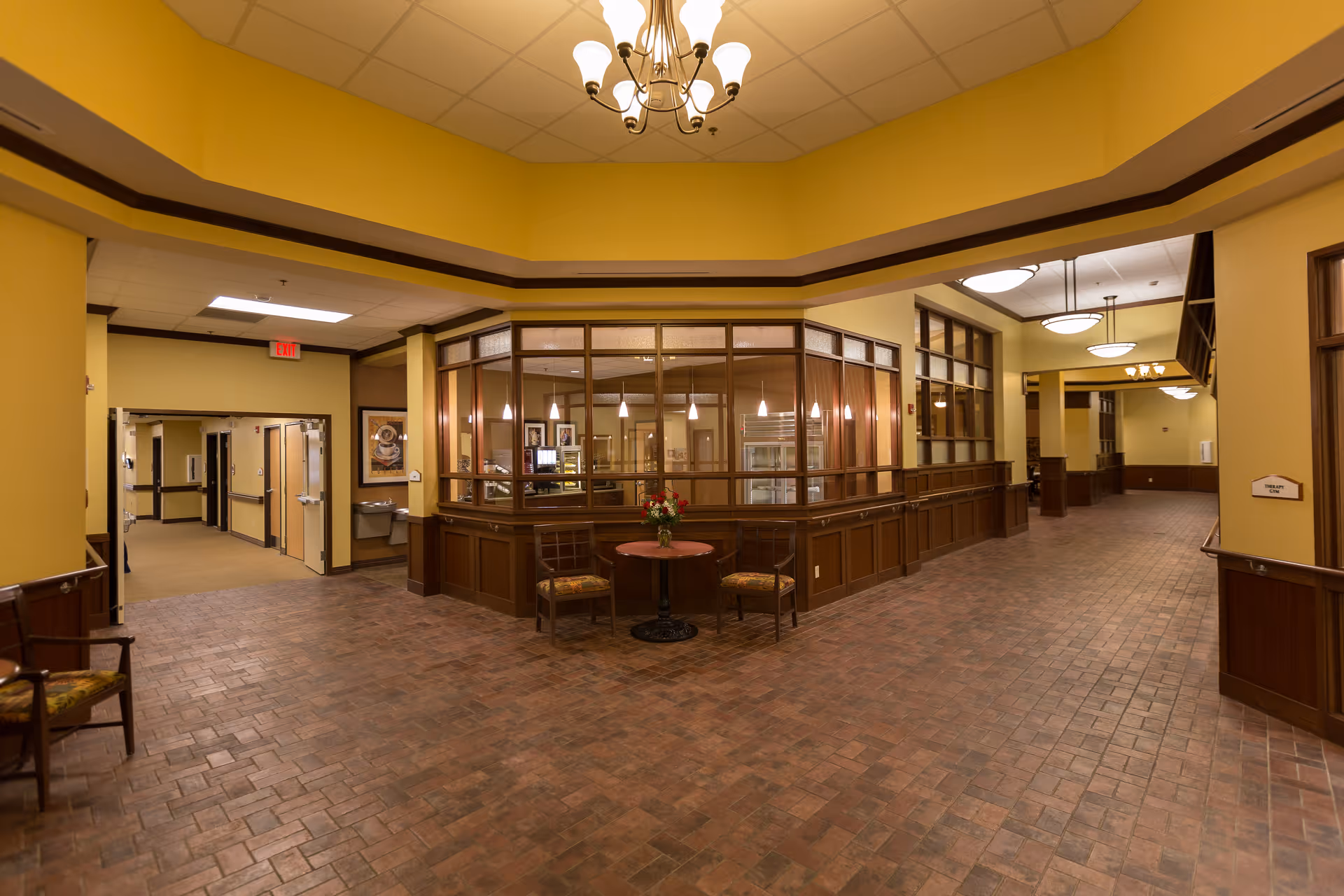 Interior view of a senior living facility hallway with yellow walls and brown trim. There is a small round table with two chairs and a flower vase in the center. The hallway has brick-patterned flooring and ceiling lights, with a glass-enclosed area that appears to be a reception or common area.