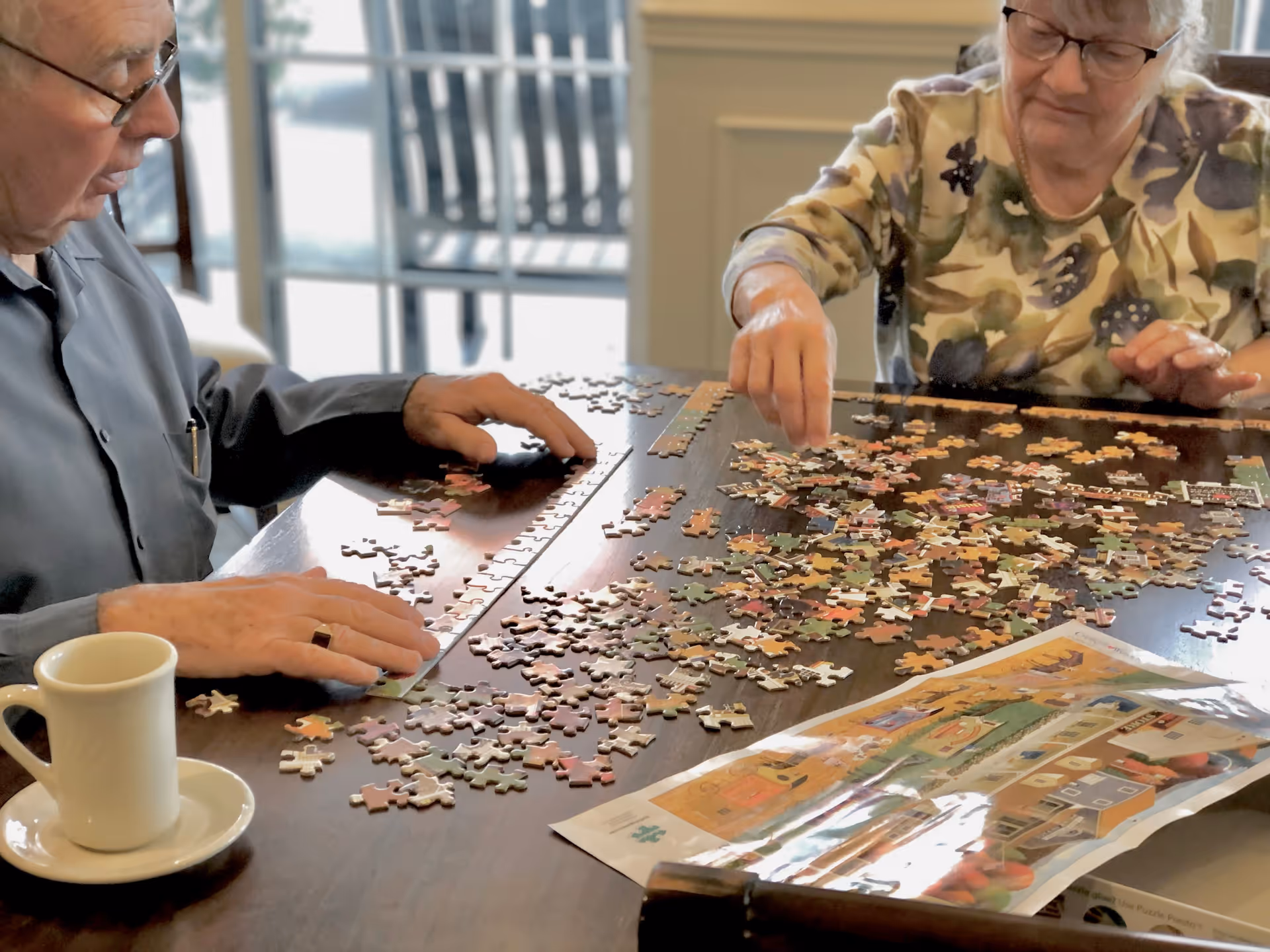 Two elderly individuals sitting at a table working together on assembling a jigsaw puzzle with many scattered pieces and a puzzle image guide on the table. A white coffee cup and saucer are also visible on the table.