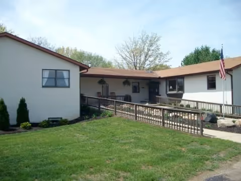 Exterior view of a single-story building with beige walls and a brown roof. There is a wheelchair-accessible ramp leading to the entrance, with railings on both sides. The building is surrounded by a well-maintained lawn and some small shrubs. An American flag is mounted on a pole near the entrance. Trees are visible in the background under a clear sky.