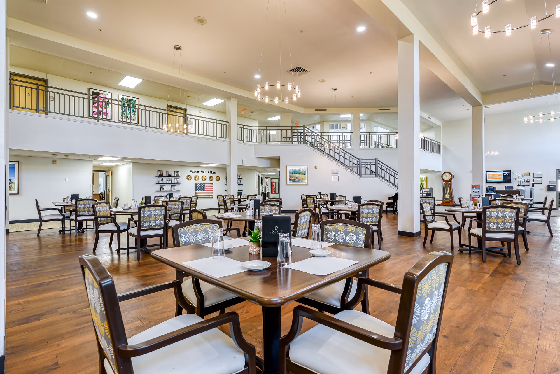 Spacious dining room in a senior living facility with multiple wooden tables and chairs arranged neatly. The room features high ceilings, modern chandeliers, and a staircase leading to an upper level with railings. The floor is wooden, and the walls are decorated with framed pictures and a Veterans Wall of Honor with an American flag.