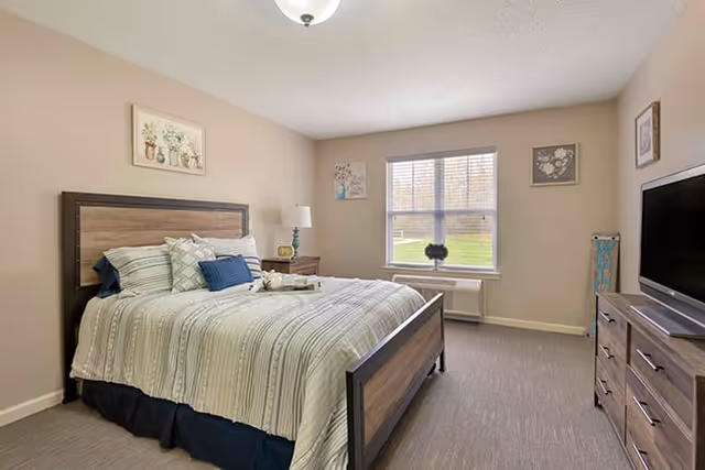 Sunny furnished bedroom with a bed, bedside table and lamp, dresser with a TV, and a window overlooking the lawn.