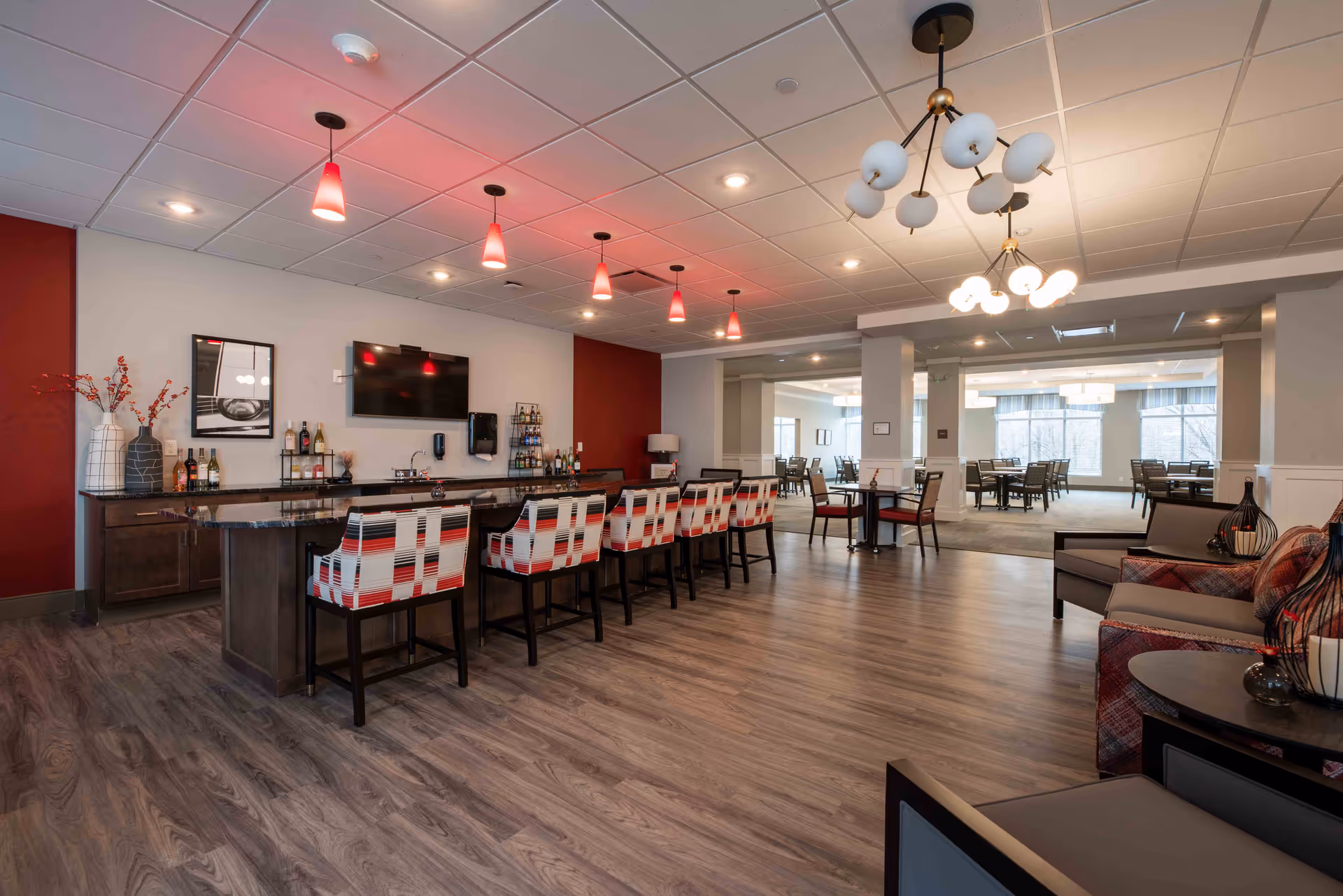 A spacious and modern common area in a senior living facility featuring a long bar with patterned chairs, pendant lights hanging from the ceiling, a wall-mounted TV, and a seating area with armchairs and tables. In the background, there is a dining area with multiple tables and chairs near large windows letting in natural light.