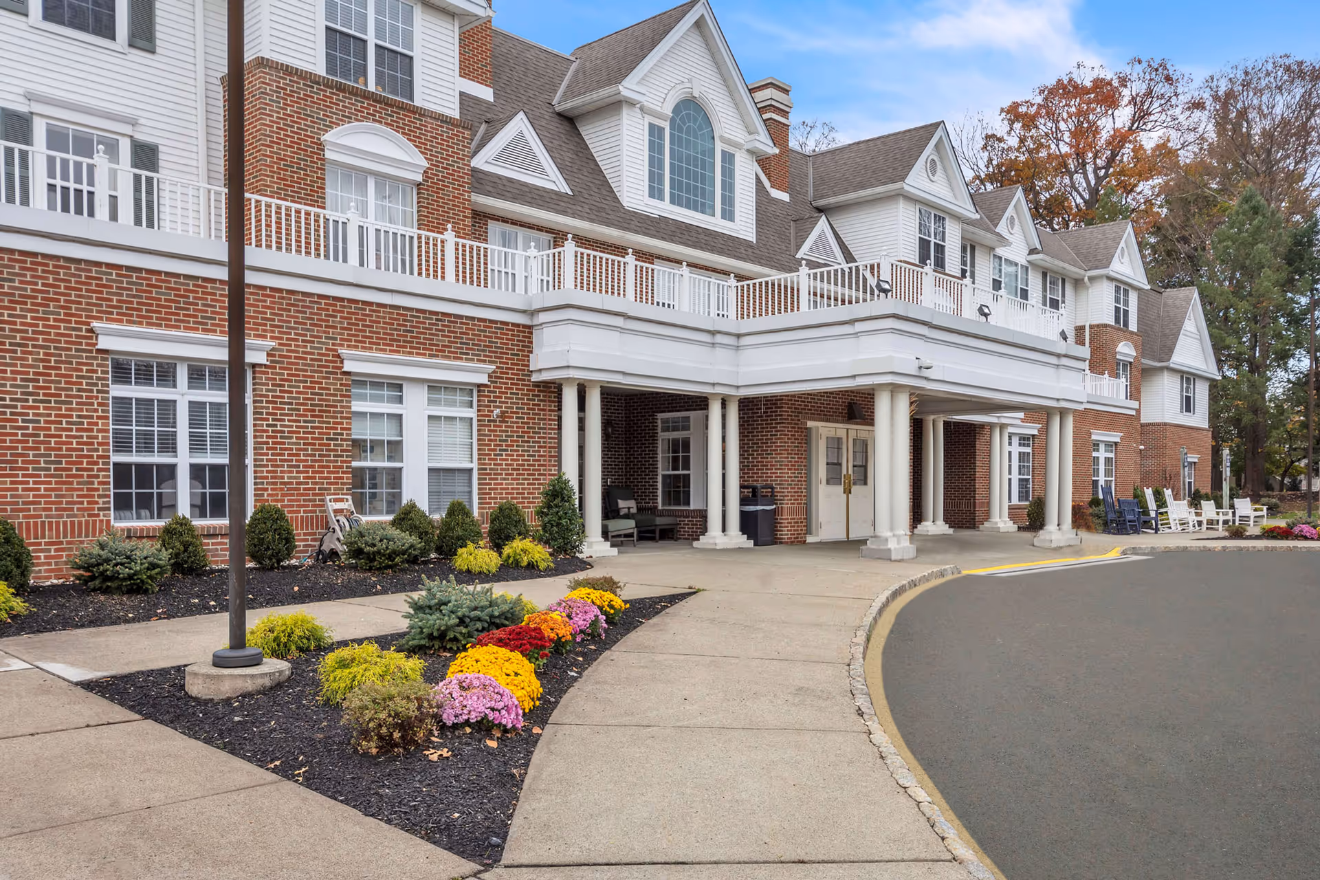 Main entrance of a red-brick senior living building with white trim, a covered drop-off portico, landscaped flower beds, and a curved driveway.